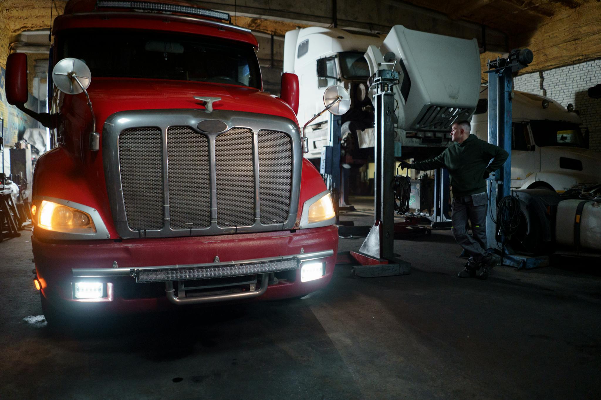 Red truck in a garage with mechanic and elevated vehicles undergoing maintenance.